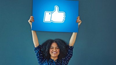 A smiling woman holds a Facebook like sign above her head.