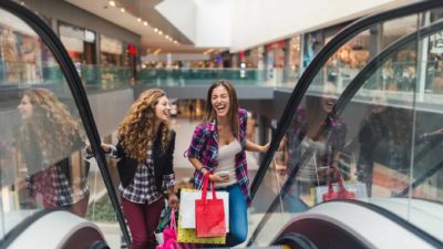 Two laughing young women hold shopping bags and ride an escalator up to another level in a Scentre Group shopping centre.
