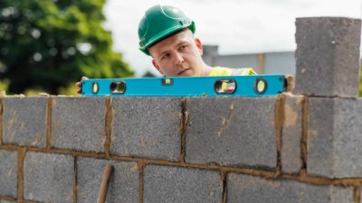 A bricklayer peers over the top of a brick wall he is laying with a level measuring tool on top and looks critically at the work he is carrying out.