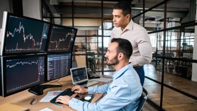 Two male ASX 200 analysts stand in an office looking at various computer screens showing share prices.