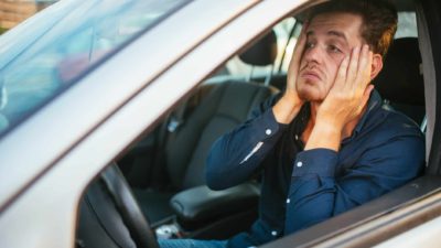 A man holds his hands to the sides of his face and pulls it down in despair as he sits at the wheel of a car that is not moving, as though in a traffic jam.