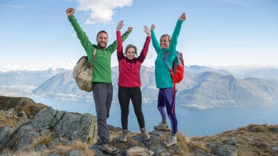 Three hikers lift their arms in jubilation as they reach a rocky peak overlooking a sensational view of water and mountains with a blue sky surrounding them.